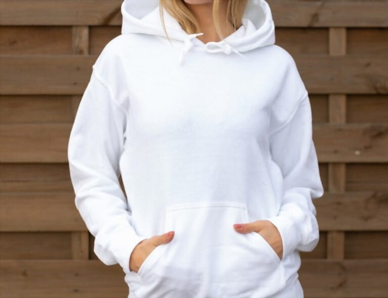 Woman is standing in white hoodie with hands in pockets in front of camera. She is posing for clothing mockup. On her hoodie is empty space for design or incription. Sweatshirt hood template