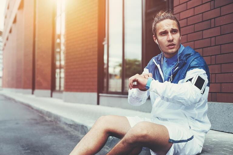 Male athlete in blue windrunner sitting on the street adjusting running program for his morning workout using smart watches on his hand