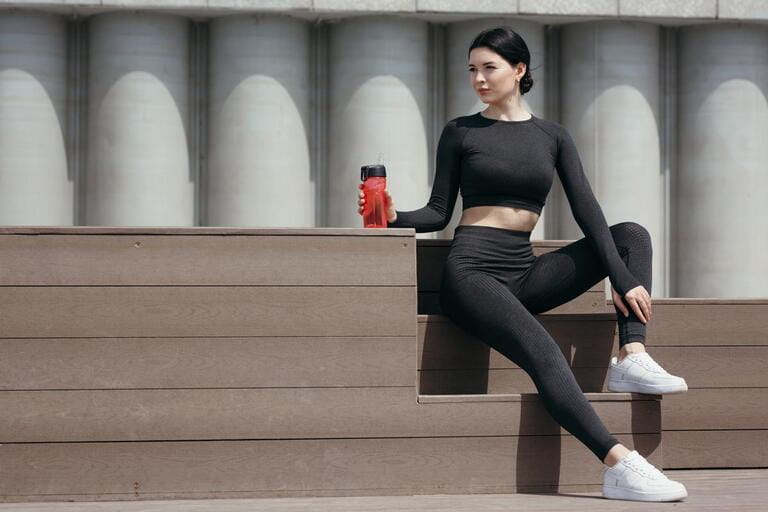 Fitness trainer in a sportwear sits on the bench and rests after jogging holding a red drinking bottle.