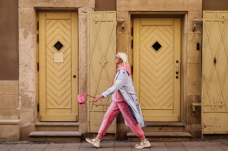 Street style, fashion: woman wearing trendy sport chic outfit walking in street of European city. Blue trench coat, sunglasses, pink hoodie, trousers, sneakers, mini bag. 