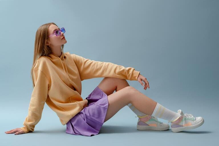 Fashionable confident girl wearing purple sunglasses, pleated mini skirt, yellow hoodie, socks, high top colorful sneakers, sitting, posing on blue background. Full-length studio portrait.